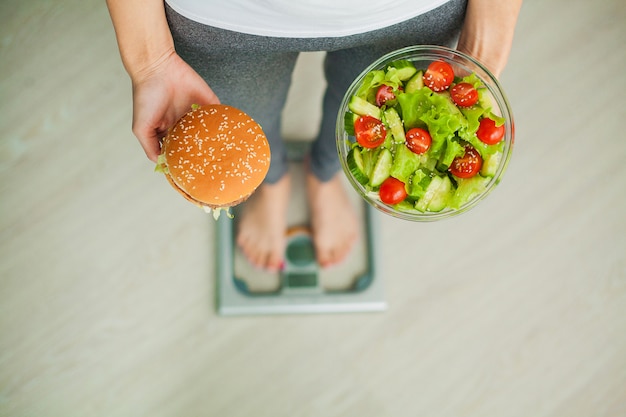 Woman measuring body weight on weighing scale holding burger and salad, sweets are unhealthy junk food, dieting, healthy eating, lifestyle, weight loss, obesity, top view Premium Photo